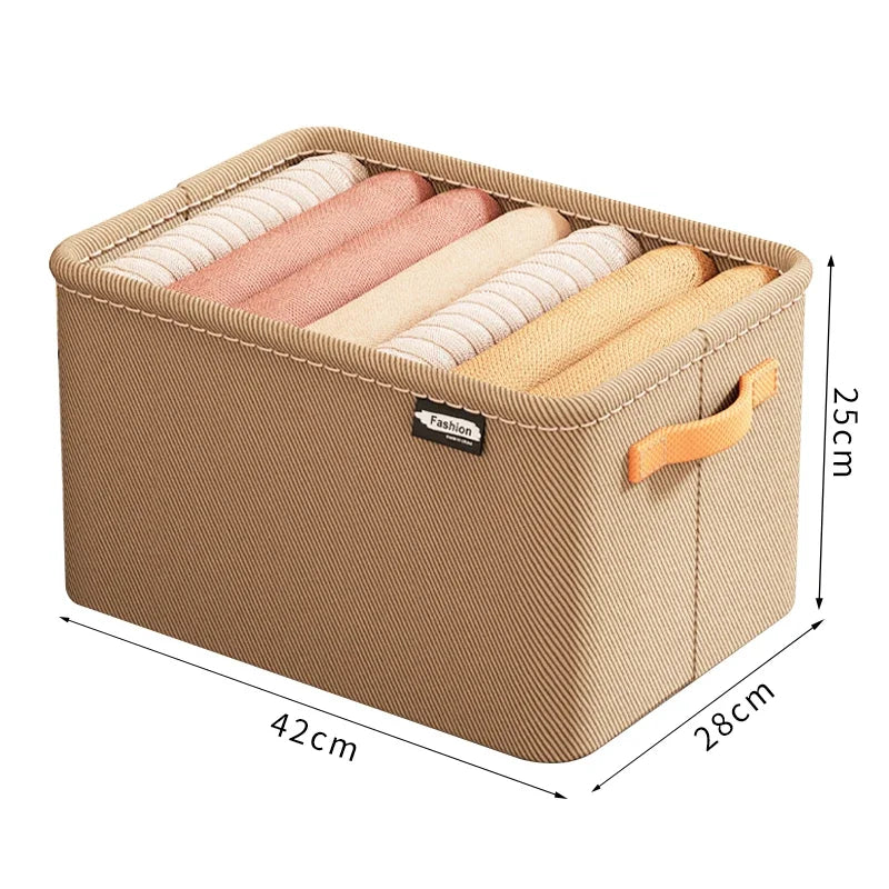 Beige stof-opbevaringskasse fyldt med oprullede tekstiler. Mål: 42x28x25 cm.