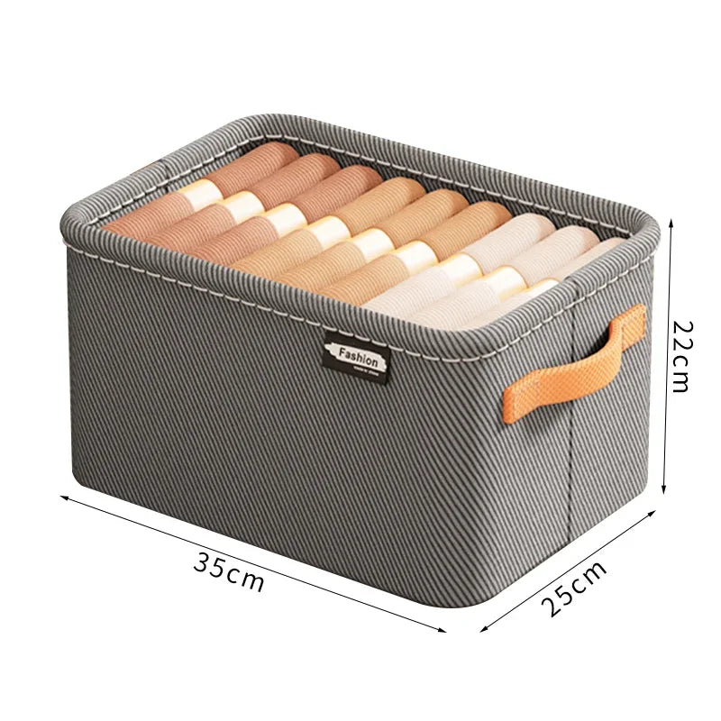 Grå rektangulær stofkurv med orange hank, hvide syninger og oprullede tekstiler indeni. Mål: 35x25x22 cm.
